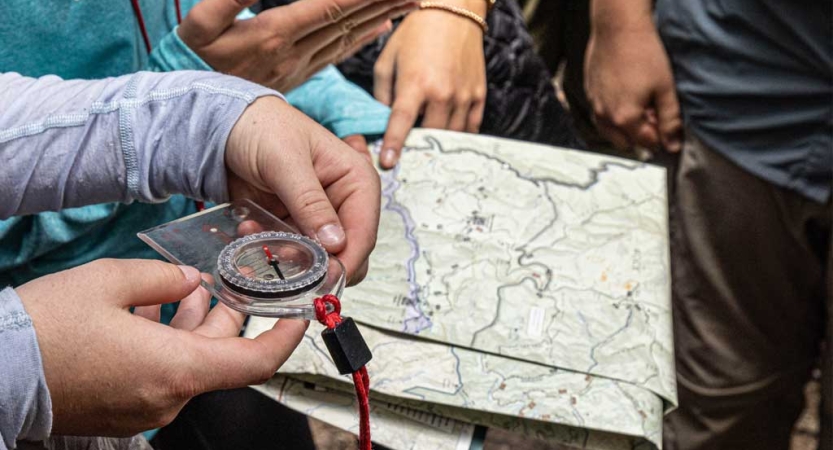 A set of hands holds a compass, while other hands are visible on a map. 